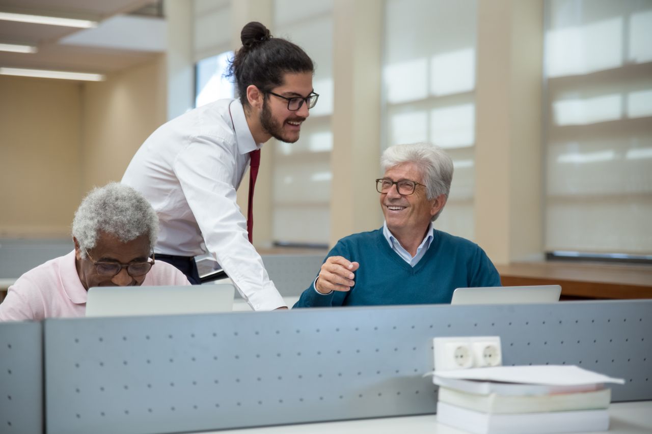 A classroom scene in Portugal where seniors learn computer skills from a young instructor. Engaged and smiling.